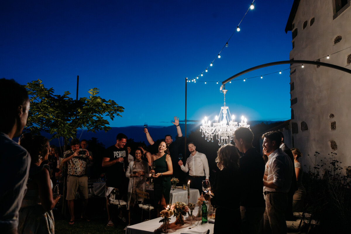Group of people celebrating outdoors at night under a chandelier with string lights.
