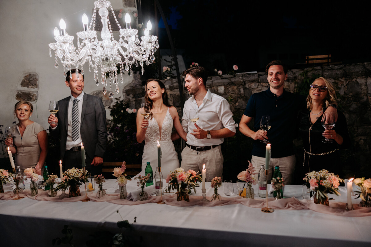 A group of six people raising glasses in a toast at a dinner table with candles and flowers.