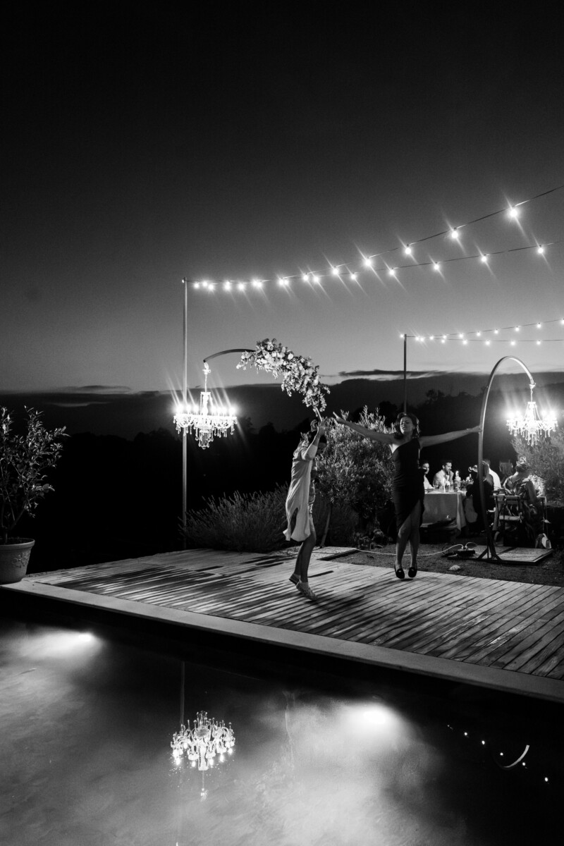 Two individuals dancing near a pool with hanging lights in a black and white setting.