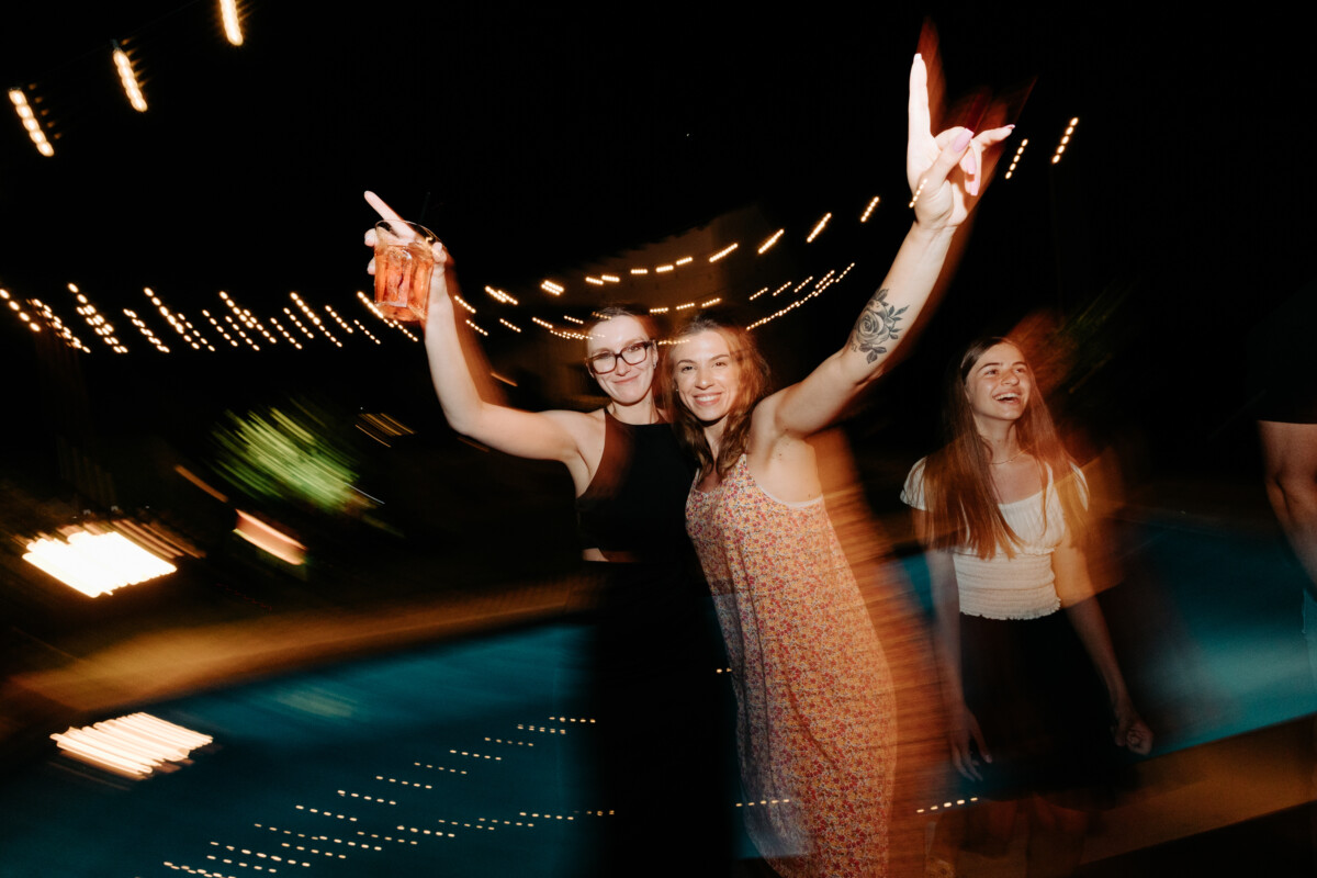 Two women raising their arms in celebration at a pool party with lights in the background.