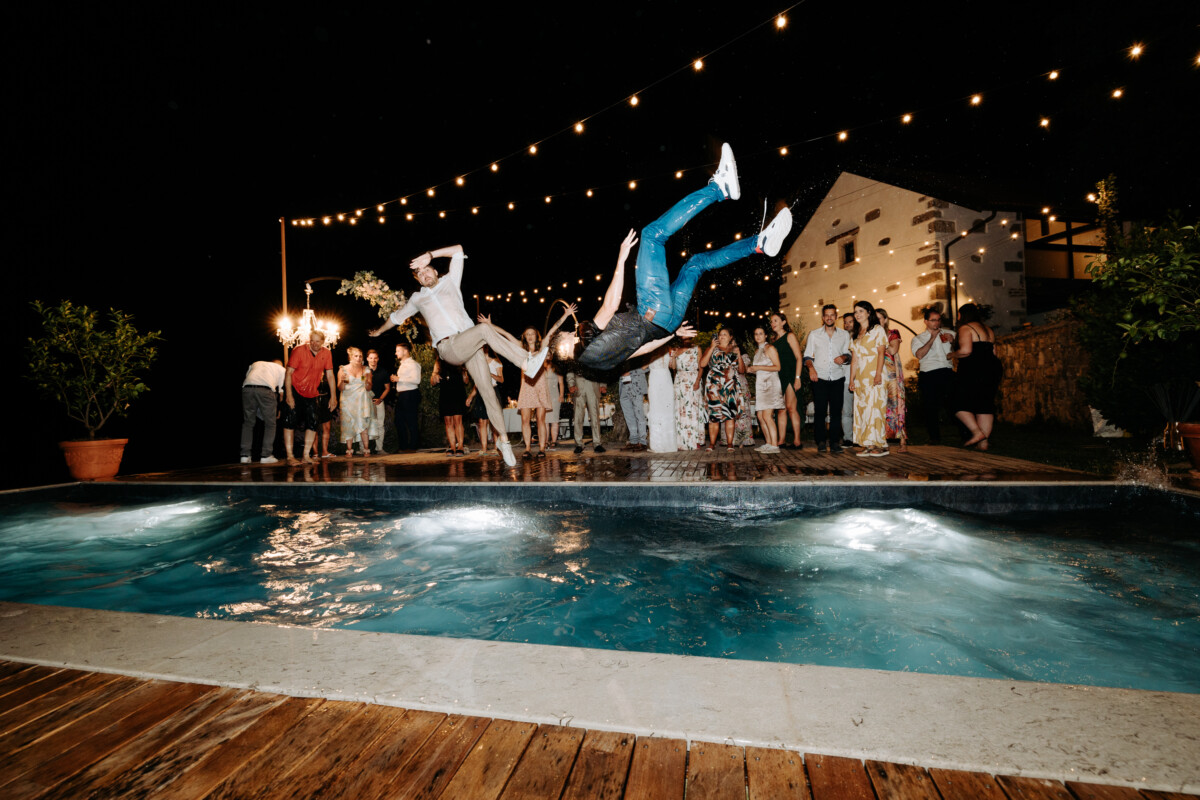 Two individuals diving into a swimming pool during a night event with string lights in the background.