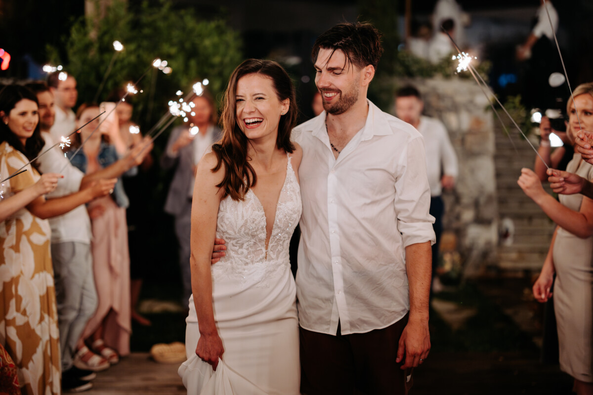 Bride in a white wedding dress and groom in a casual shirt smiling while surrounded by guests holding sparklers.