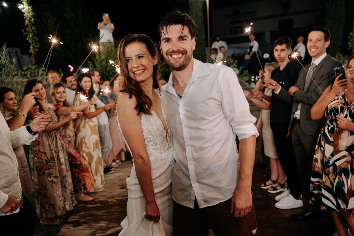 Bride and groom smiling together during a wedding celebration surrounded by guests holding sparklers.