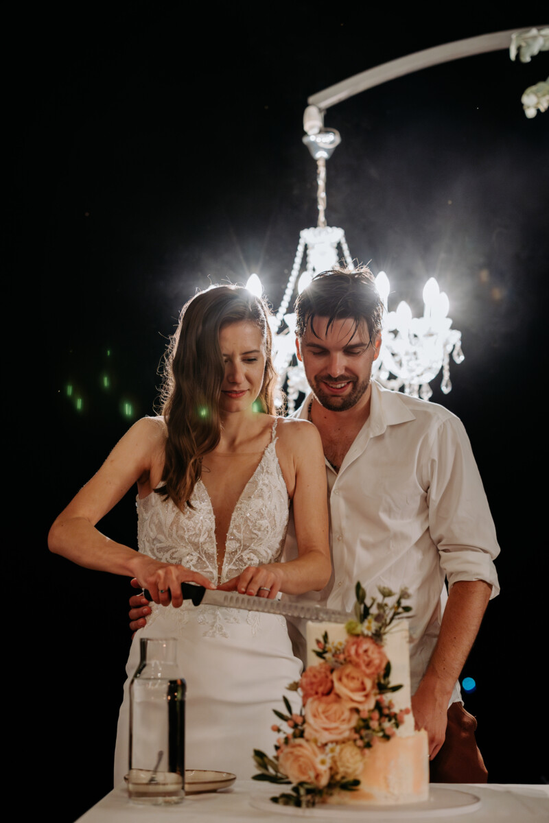 Bride and groom cutting a wedding cake together under a chandelier.