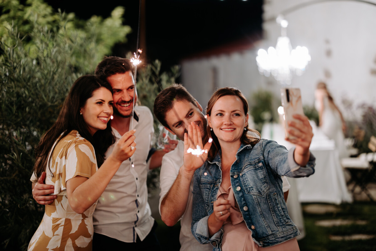 Four friends taking a selfie while holding sparklers at an outdoor gathering.