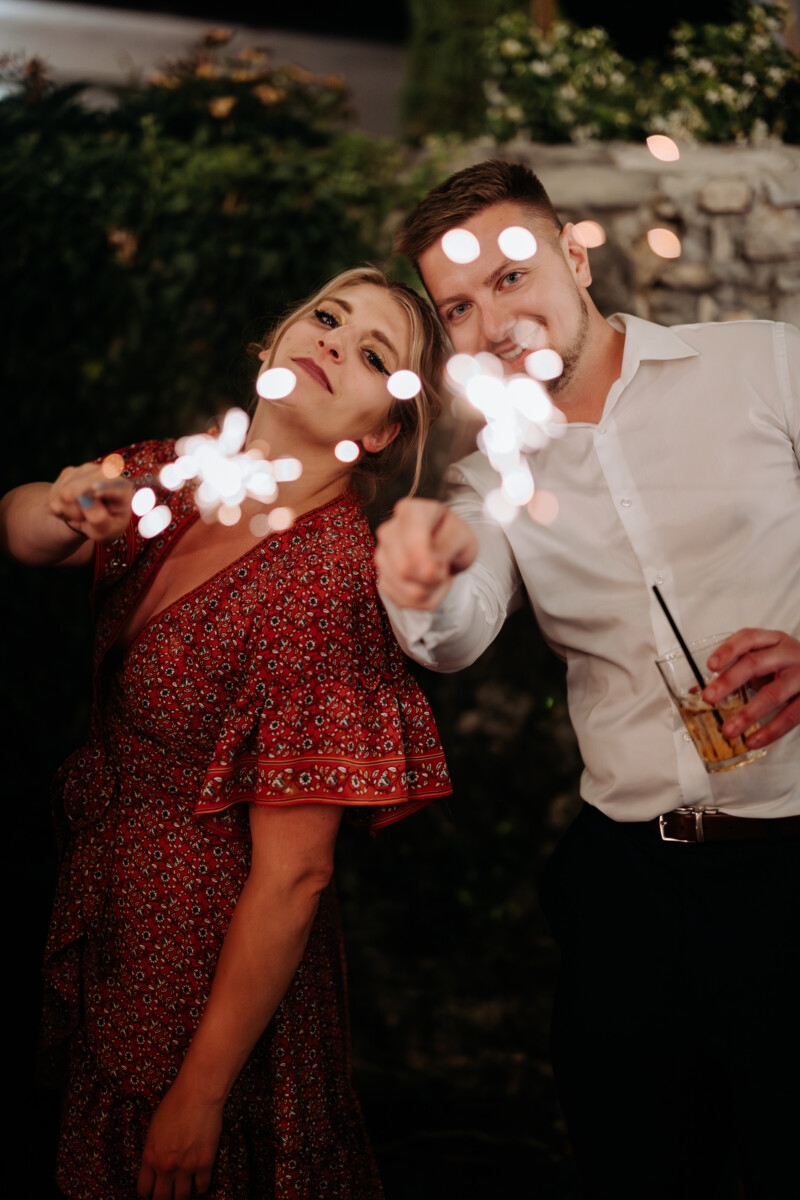 A couple holding sparklers, with lights glowing in the dark, celebrating an event outdoors.
