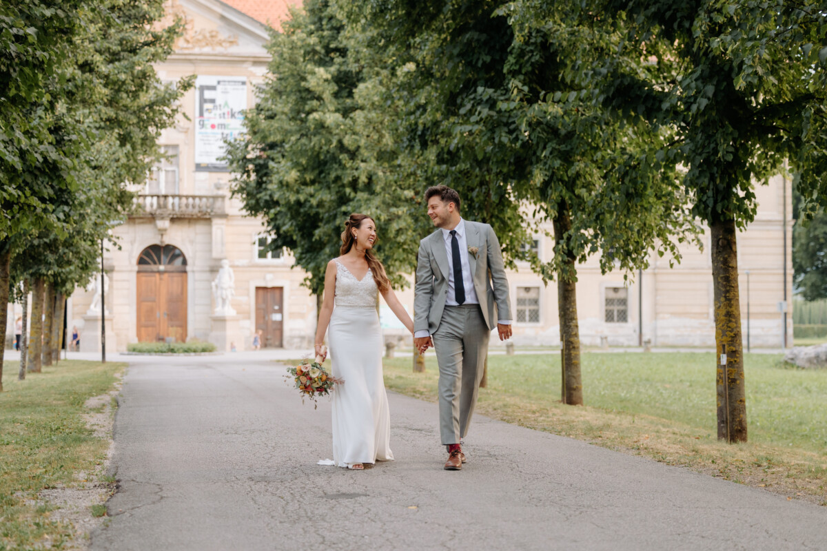 A bride in a white dress and a groom in a gray suit walking hand in hand on a path.